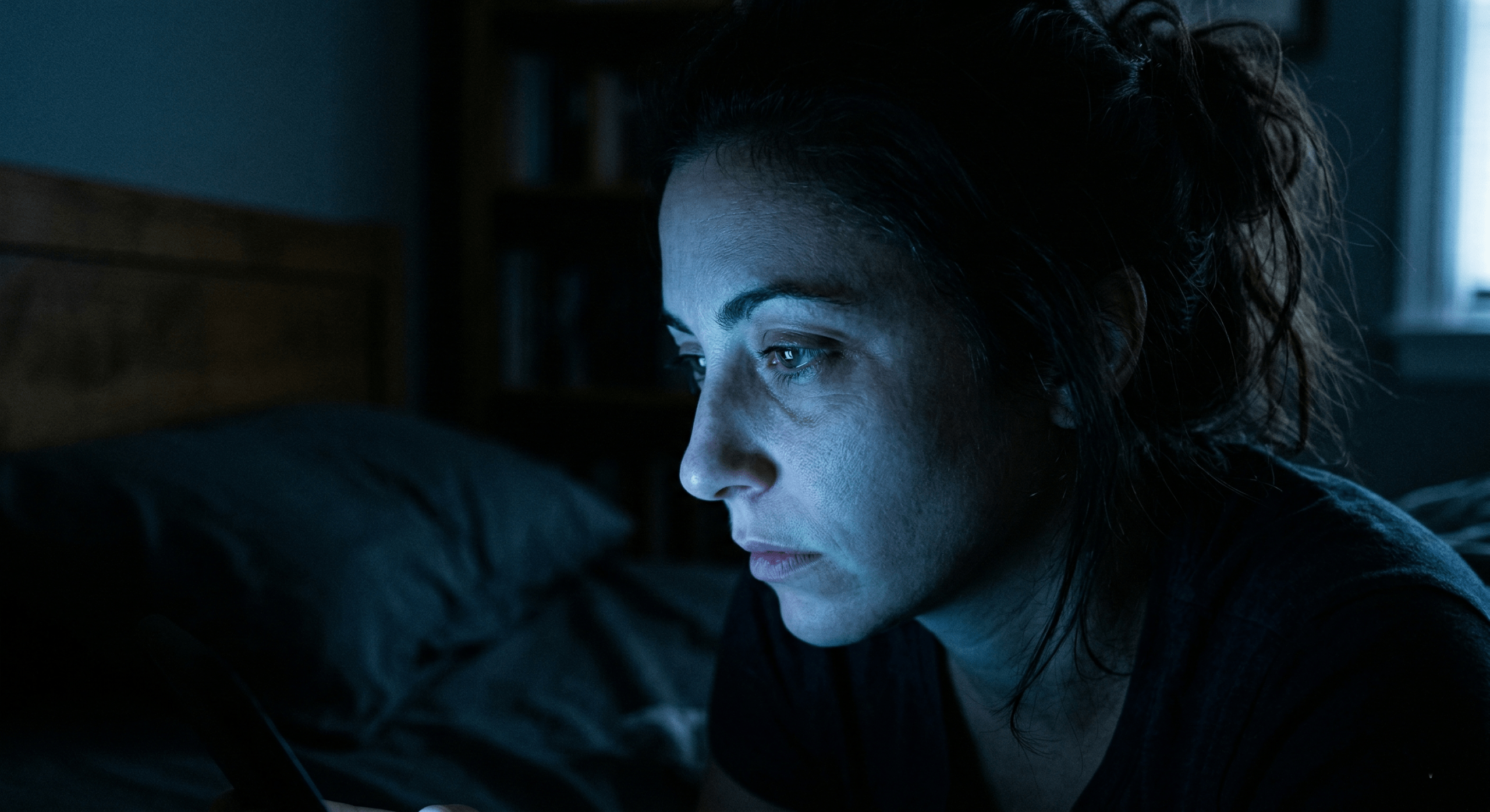 Close-up photograph of a person's tired profile illuminated by harsh blue light in a dark bedroom at night, illustrating sleep disruption.
